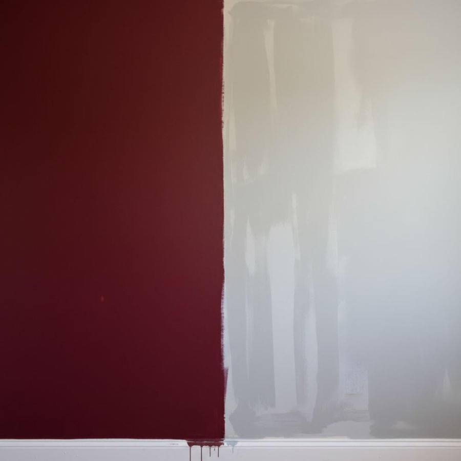 A close-up shot of a wall with two distinct colors: a rich burgundy section freshly painted on one side, while the other side of the wall remains untouched and waiting for its second coat.